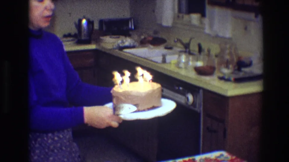 A closer 8mm frame shows the woman with the lit birthday cake beside kitchen glassware, a sink, and countertop items.