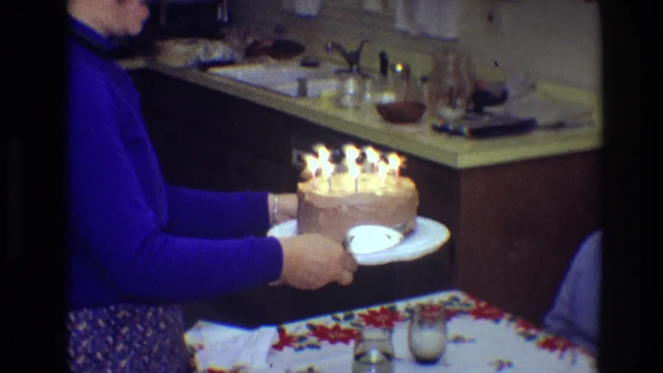 The woman holds the candlelit birthday cake above a table with a glass and patterned cloth in the 1968 Toledo kitchen scene.