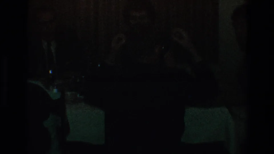 Very dark 1968 Toledo home-movie frame showing silhouetted people seated around a dining table, with glassware faintly visible.