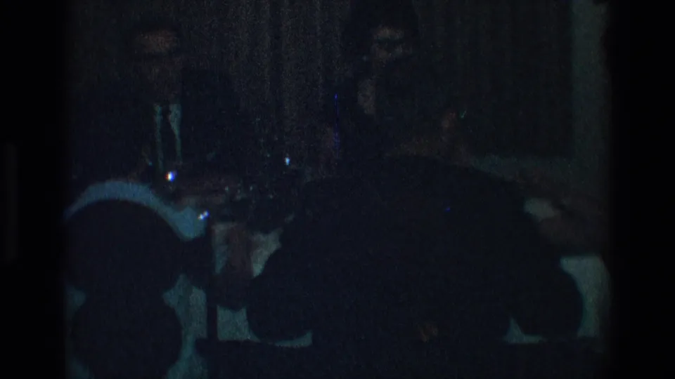 Dark, grainy 8mm frame from Toledo in 1968 showing several people seated at a dinner table with glassware and dishes in low light.