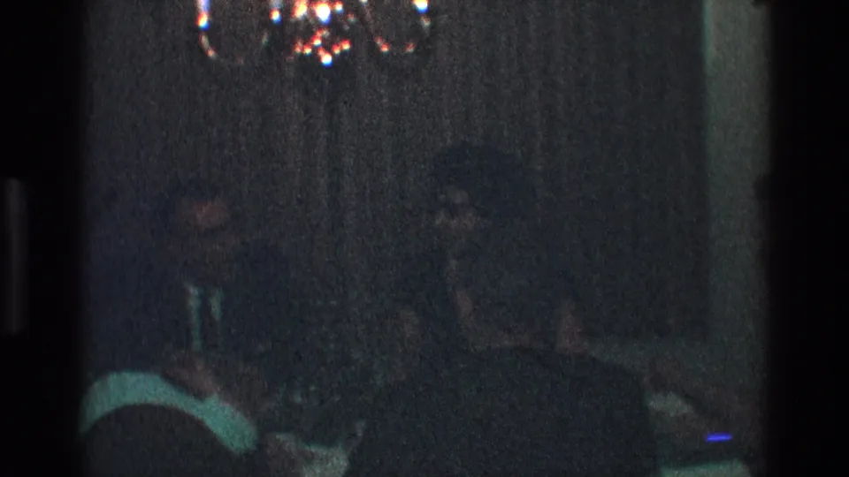Dim 8mm frame from a 1968 Toledo dinner party showing seated adults, tableware, curtains, and a chandelier overhead.