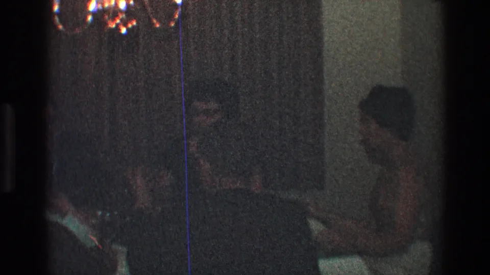 Late-1960s Toledo home-movie frame with a woman seated at right and other dinner guests partly obscured in a dark interior.