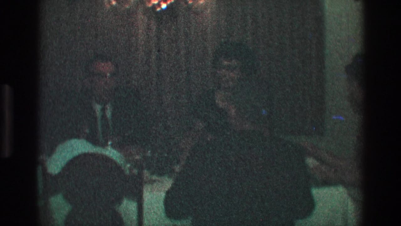 Adults in suits sit at a table with curtains behind them and a chair in front.