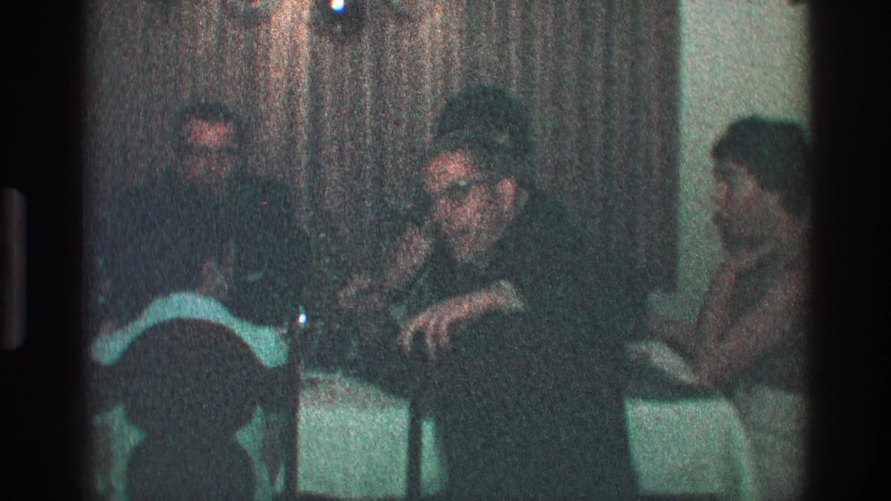 Adults seated around a dim dinner table in a grainy interior frame.