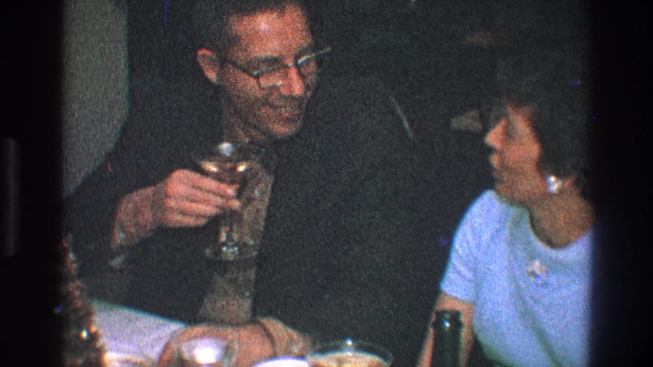 Man with glasses holds a drink beside a seated woman at a table.