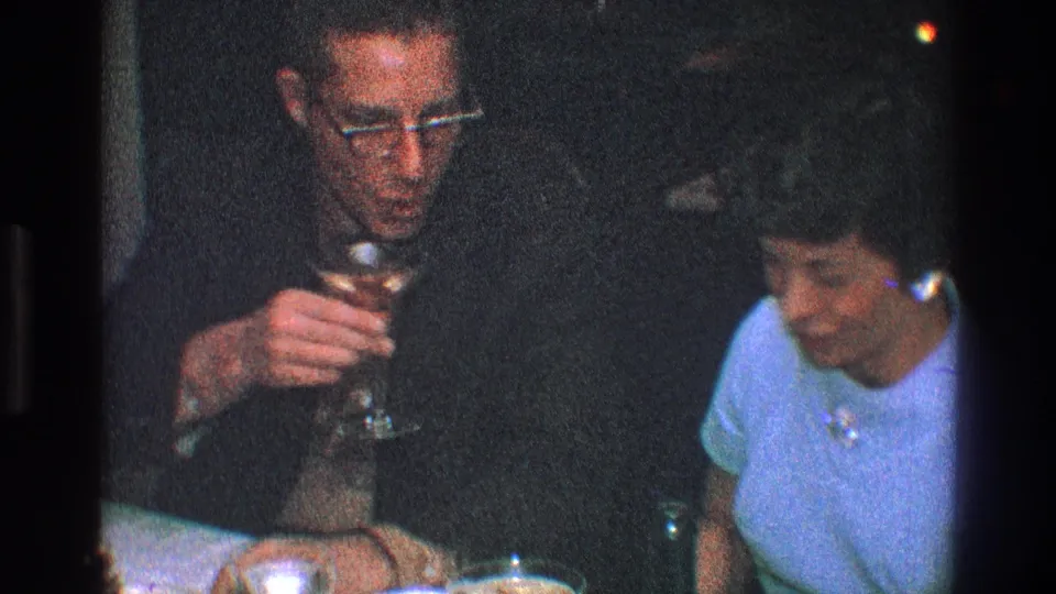 1968 Toledo home-movie frame showing a man raising a drink near a woman seated at a dim dinner table.