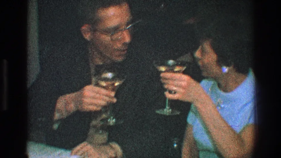 The seated couple holds martini glasses in low light, with the man looking down and the woman turning toward him.
