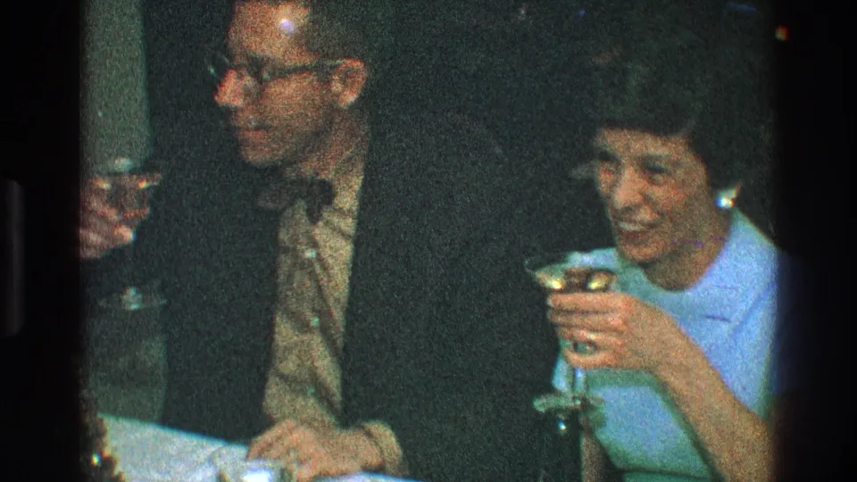The woman smiles while holding her martini glass as the man sits beside her in formal clothing at the dinner table.