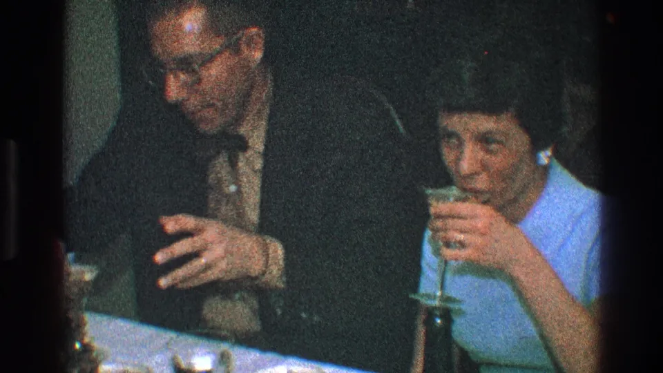 The woman sips from her martini glass while the man leans nearby, framed tightly in the dim 8mm dinner scene.