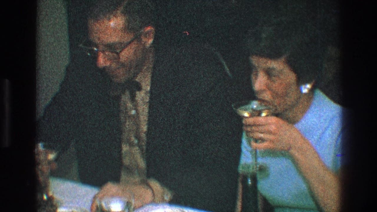 Man in glasses and woman in a light blue dress sit at a table with drinks.