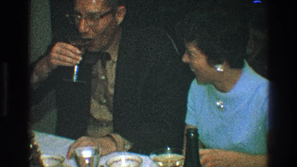 The man sips from a small stemmed glass as the woman turns toward him during a dim 1968 Toledo dinner scene.