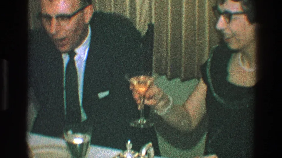 A man in a dark suit and a woman in a dark dress sit before curtains as the woman lifts a cocktail glass during the Toledo, Ohio dinner scene.