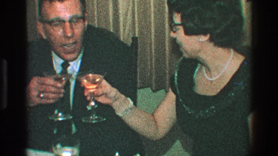 A suited man and a woman in a dark dress raise cocktail glasses toward each other at a dimly lit table in the 1968 Toledo home-movie footage.