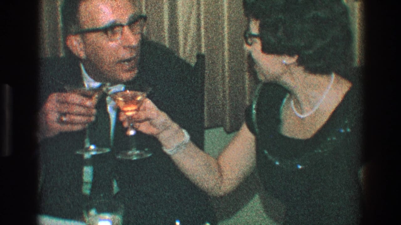In 1968 Toledo home-movie footage, a suited man with glasses and a woman in a dark dress with pearls hold stemmed drinks while seated at a table.