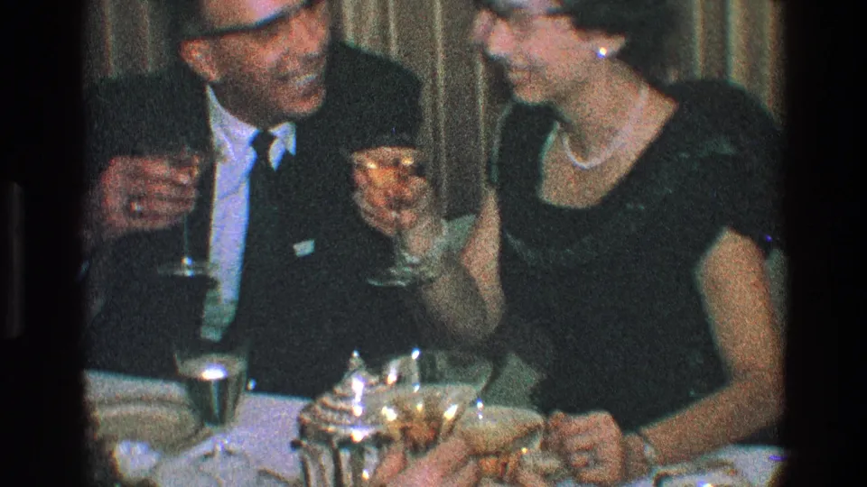 After the kiss, the man and woman sit close together at a dinner table with stemmed drinks, plates, and a reflective teapot or coffee pot visible.