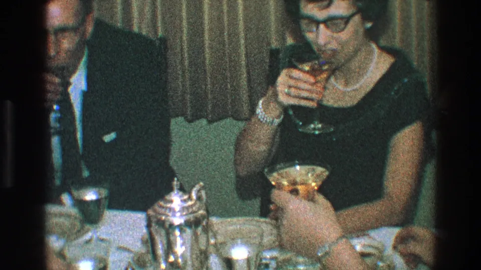 The woman in a dark dress and pearl necklace drinks from a stemmed glass at the table, with the suited man and another hand with a cocktail partly visible nearby.
