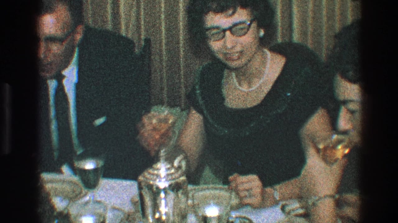 Woman in dark dress smiles at a table while adults hold drinks nearby.