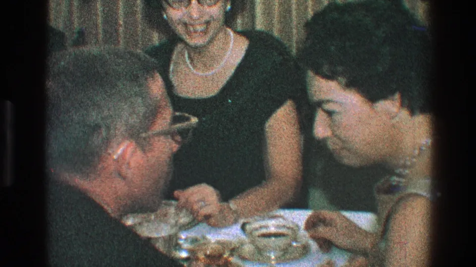 Adults lean in and talk around a dinner table, with a woman in pearls and other guests framed closely by the handheld 8mm camera.