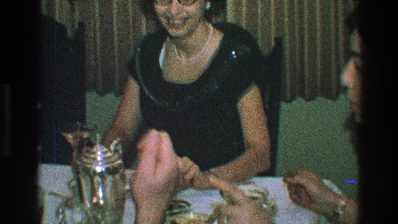 Woman in a black dress and pearls seated at a table while guests hold drinks nearby.