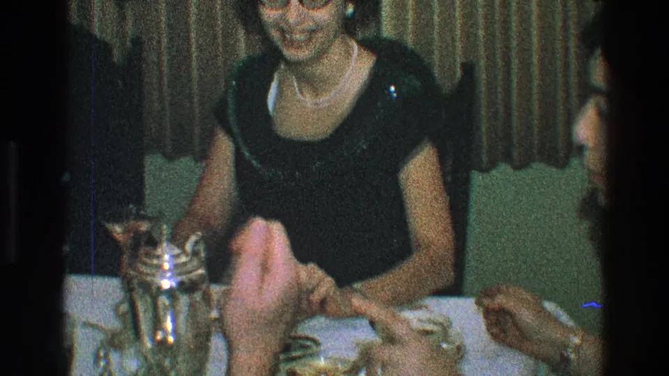 1968 Toledo home-movie frame showing a woman in a black dress and pearl necklace at a dinner table with stemware and a silver pitcher.