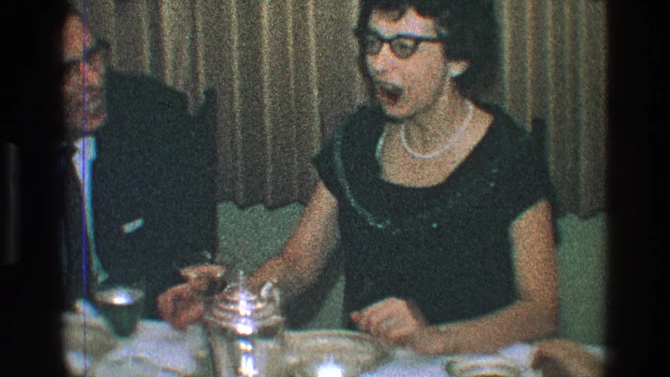1968 Toledo home-movie frame of a woman in glasses and pearls laughing near a suited man, with table settings and curtains behind them.