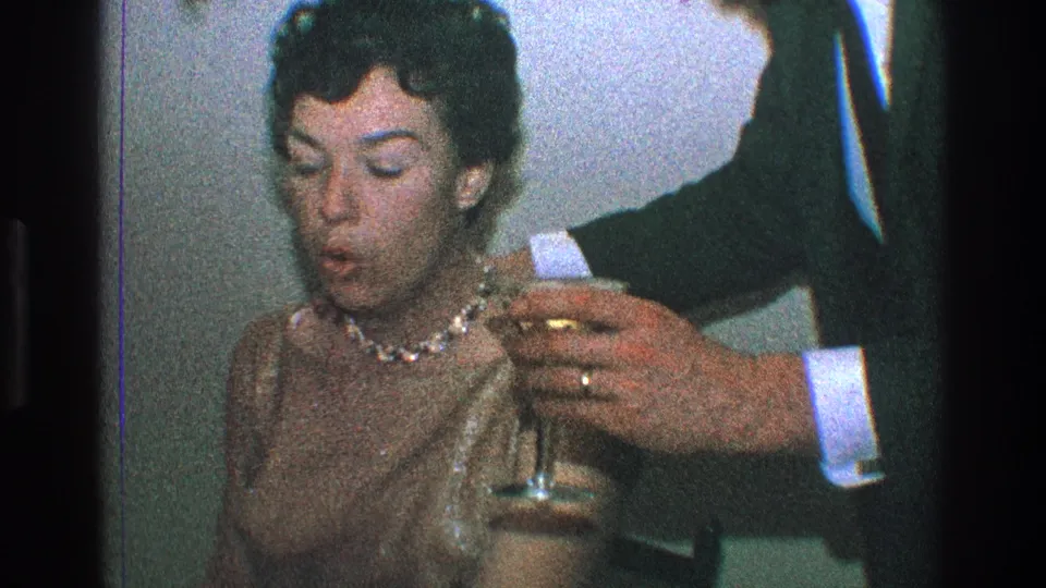 1968 Toledo home-movie frame showing a woman in a sequined dress and pearls seated beside a suited man holding a drink.