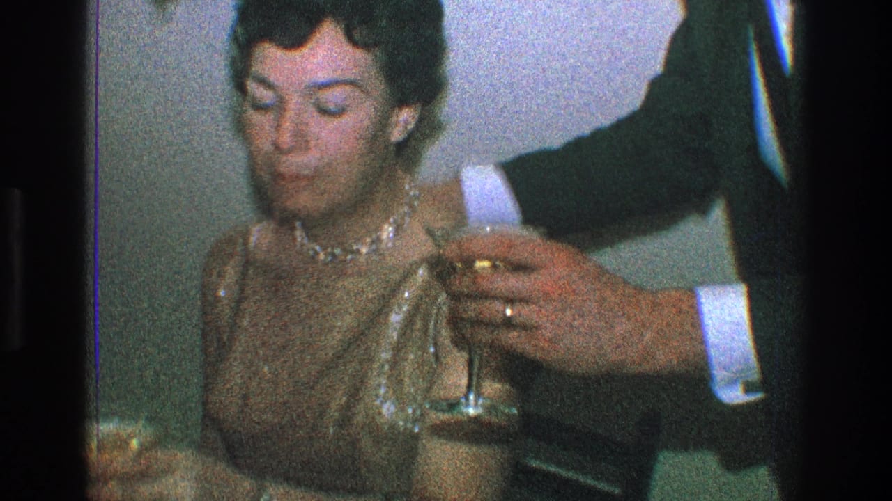 In a 1968 Toledo, Ohio home-movie party scene, a seated woman in a shimmering sleeveless dress looks down while a suited man reaches in with a stemmed glass.