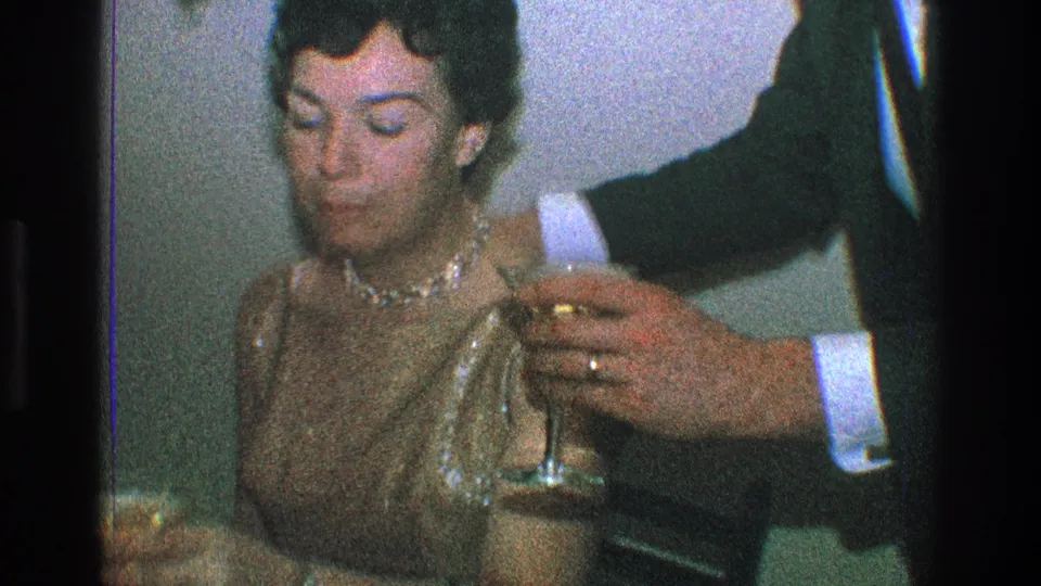 In a 1968 Toledo, Ohio home-movie party scene, a seated woman in a shimmering sleeveless dress looks down while a suited man reaches in with a stemmed glass.