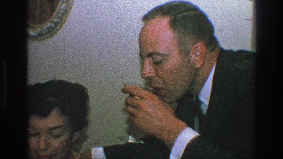 A suited man takes a drink from a small glass beside the seated woman in the private indoor party setting documented on 8mm film.