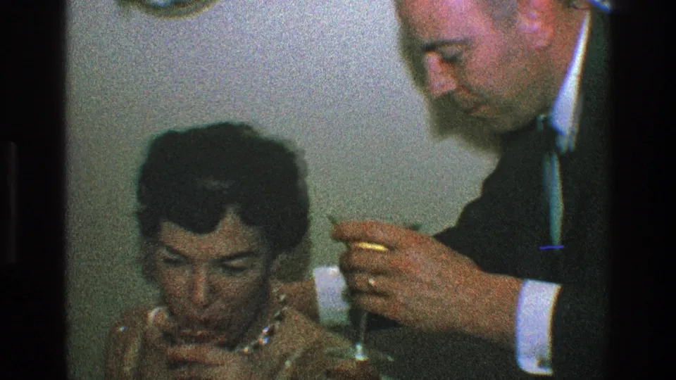 The woman looks down with a glass near her hand as the suited man remains close, continuing the intimate party toast moment in the 1968 Toledo home movie.