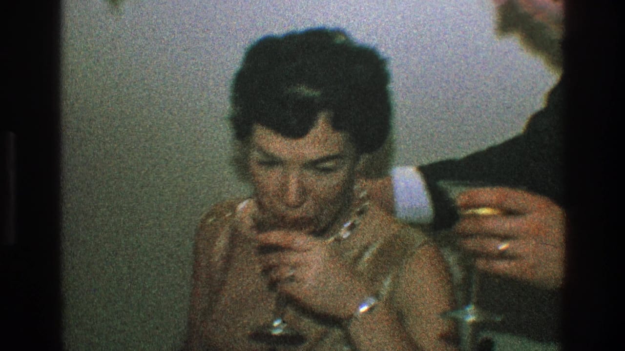 Woman in a gold dress sits with a wine glass as a suited man stands close beside her.