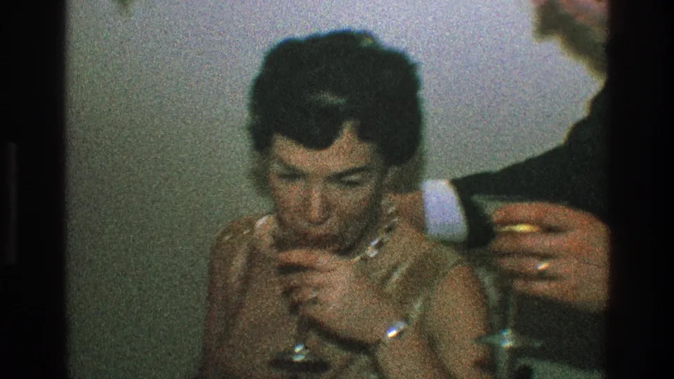 In 1968 Toledo home-movie footage, a woman in a shimmering dress sits at a table while a man in a dark suit holds a stemmed glass nearby.