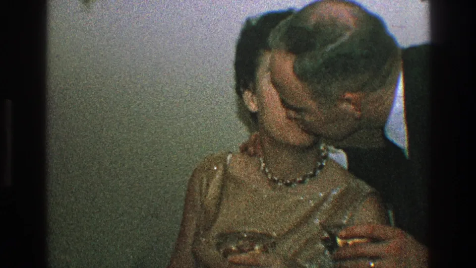 A man in a dark suit kisses a seated woman in a gold dress as she holds a stemmed glass in this 1968 Toledo home-movie frame.