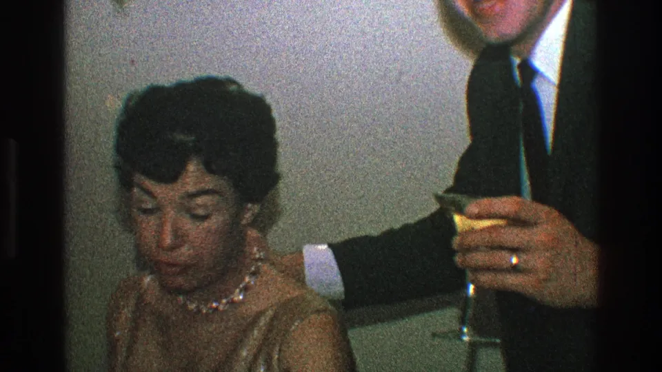 In the Toledo 1968 private interior, the woman looks downward as a suited man beside her holds a stemmed glass near the table.