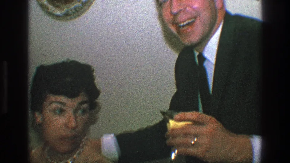 In a 1968 Toledo home-movie frame, a woman in a beaded necklace and a suited man laugh close to the camera with drinks visible.