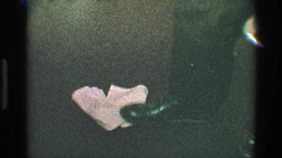 1968 Toledo home-movie frame showing a low close view of dancing legs and shoes near a crumpled napkin on dark carpet.