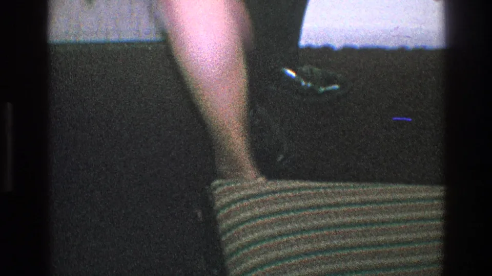 1968 Toledo home-movie frame showing cropped adults in dress clothing indoors beside a striped seat or cushion.