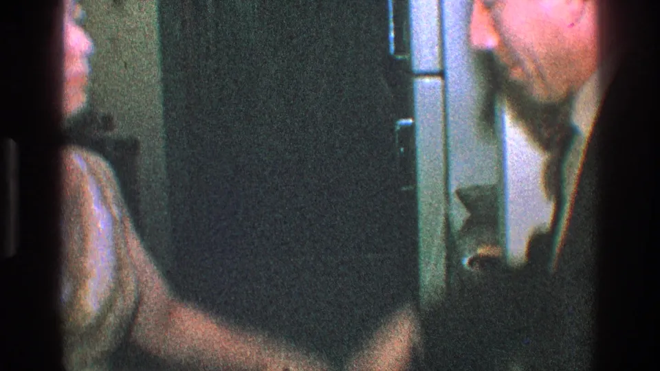 Cropped 1968 Toledo frame of a suited man leaning near a woman in a private interior, with soft focus and dark film borders.