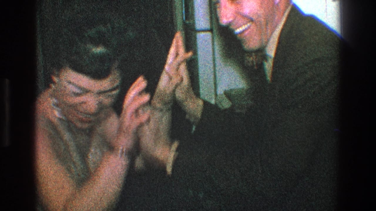 In supported 1968 Toledo home-movie footage, a woman in a sparkly top covers her face as a suited man smiles during a playful indoor exchange.