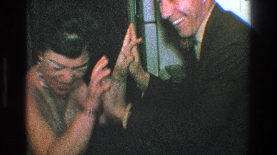 In supported 1968 Toledo home-movie footage, a woman in a sparkly top covers her face as a suited man smiles during a playful indoor exchange.