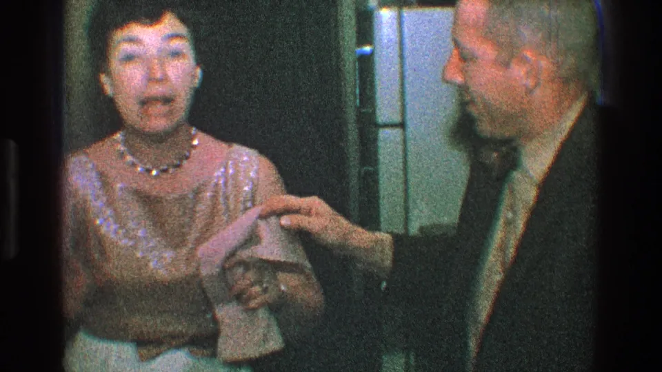 In 1968 Toledo home-movie footage, a woman in a gold dress and pearl necklace reacts while a suited man stands beside her indoors.