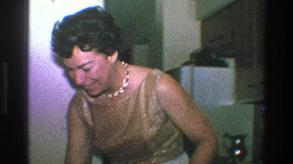A woman in a gold dress and pearls looks down inside a Toledo home during the 1968 party footage.