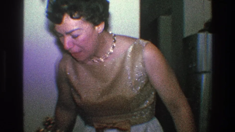 The 1968 Toledo home movie shows the woman leaning forward in a gold dress, with a refrigerator and interior wall behind her.