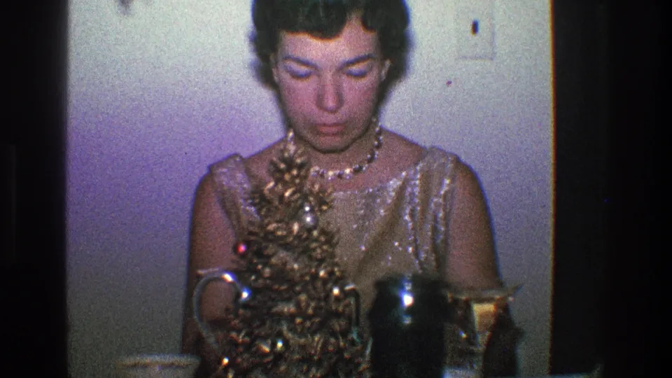 The woman looks down at the tabletop Christmas decoration, with reflective pitcher and cup visible in the foreground.