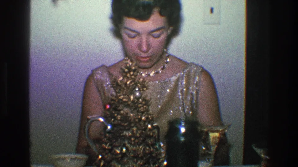 In this 1968 Toledo home-movie frame, a woman in a shimmering gold dress looks down at a table with a small decorated Christmas tree and glassware.