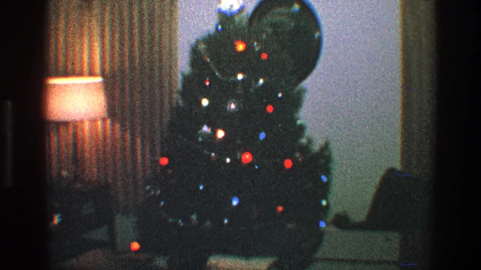 1968 Toledo home-movie frame with a softly lit Christmas tree, warm lamp, patterned curtains, and dark room furnishings.