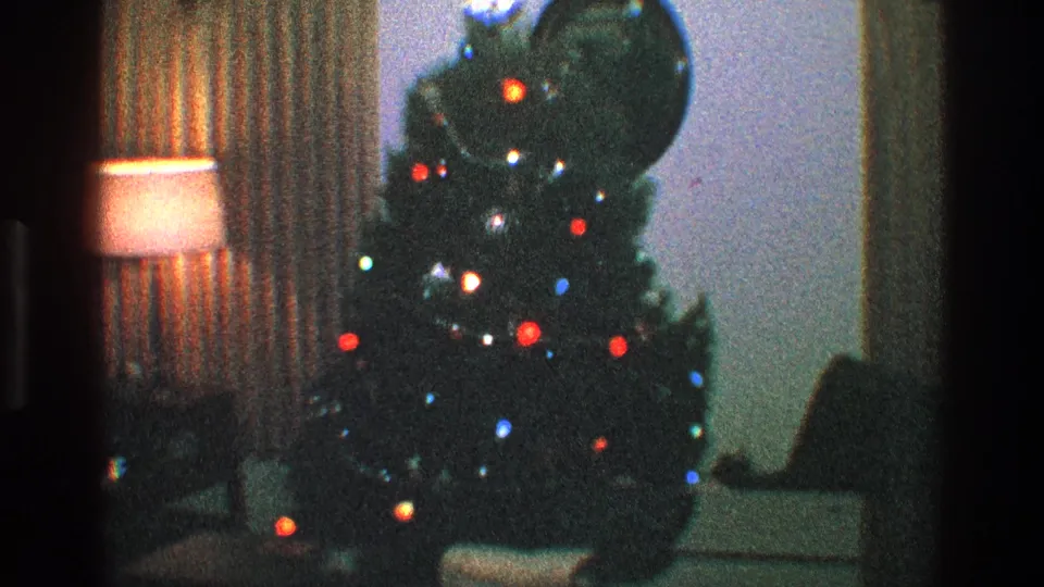 1968 Toledo home-movie frame showing a lit Christmas tree near a warm lamp, curtains, and seating inside a private living room.
