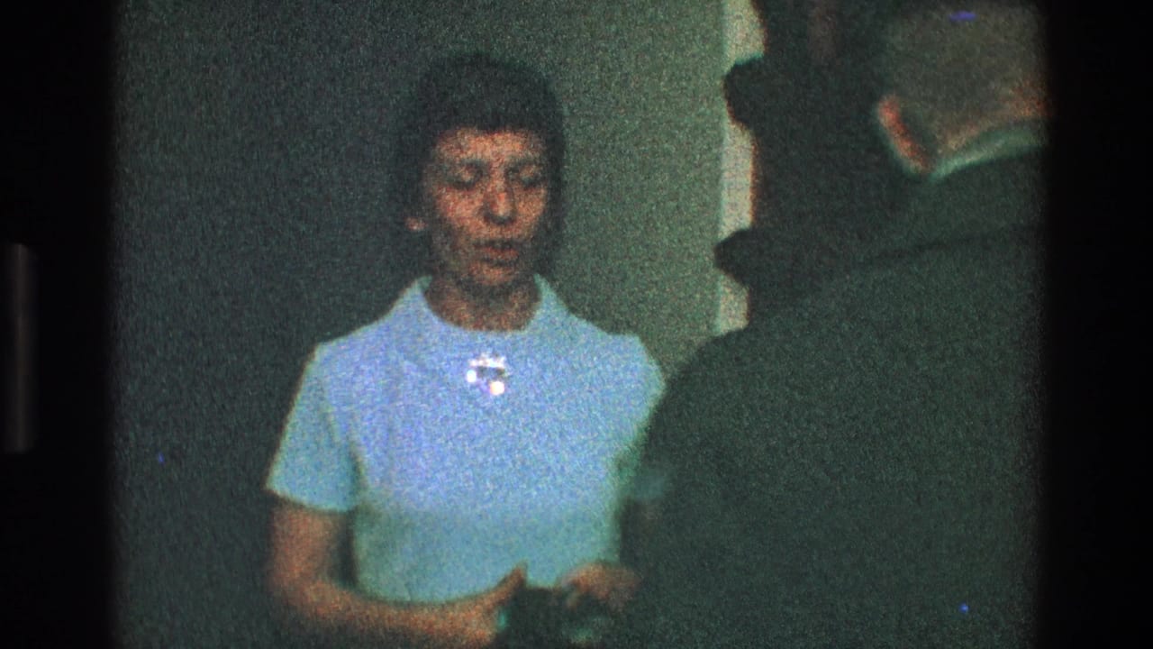 Woman in a light blue dress faces a man in a dark coat near an interior doorway.