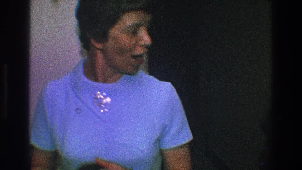 The woman in a light blue short-sleeved dress turns to the side, her brooch catching light in the grainy 1968 Toledo home movie.