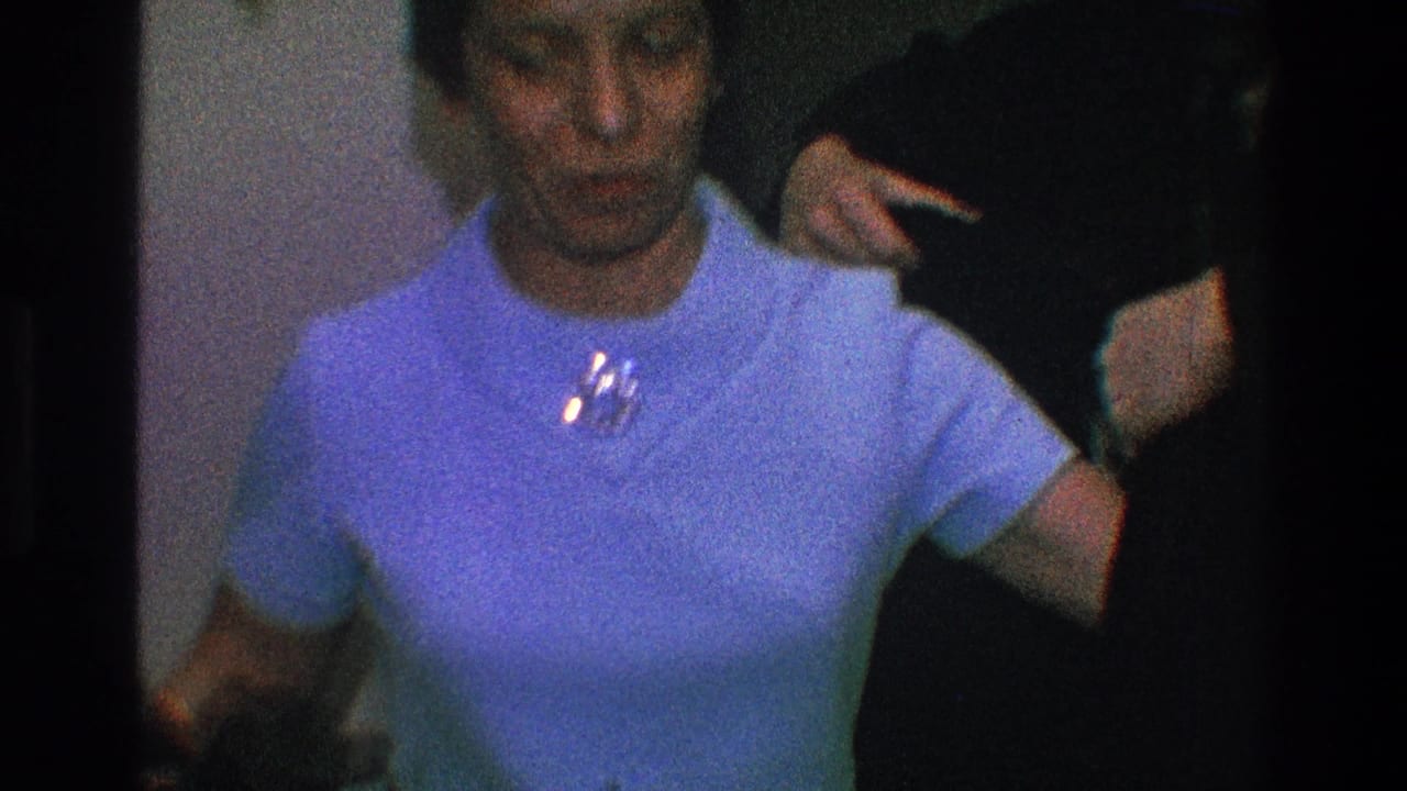 In 1968 Toledo home-movie footage, a woman in a light blue dress and brooch stands while someone helps position a dark coat behind her.